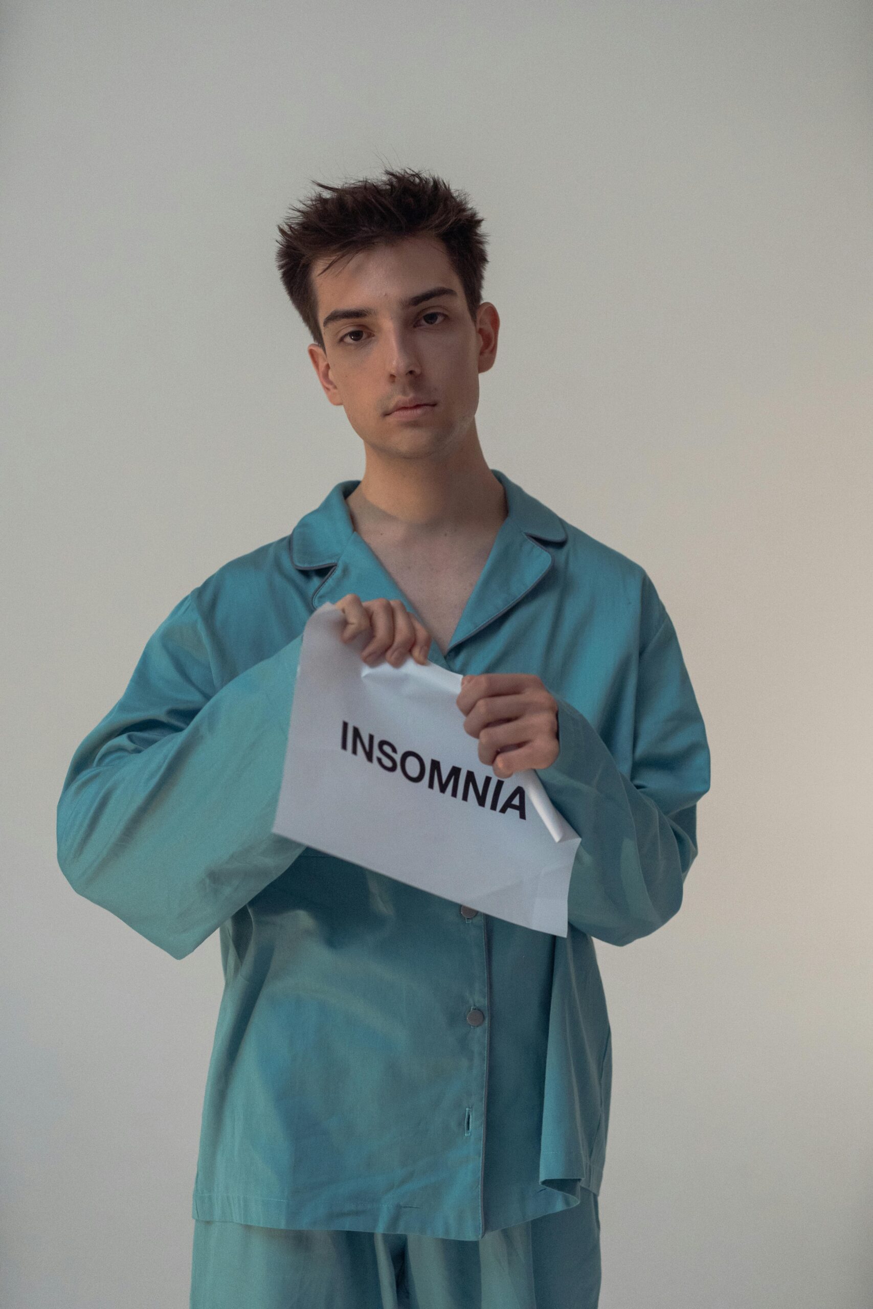 Tearing Down the Night: A Defiant Act to Banish Insomnia | Calmly Rooted A young person with dark curly hair and a neutral expression, wearing teal satin pajamas, looks directly at the camera while tearing a white piece of paper in half. The paper is printed with the word "INSOMNIA" in black capital letters.