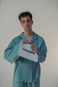 Tearing Down the Night: A Defiant Act to Banish Insomnia | Calmly Rooted A young person with dark curly hair and a neutral expression, wearing teal satin pajamas, looks directly at the camera while tearing a white piece of paper in half. The paper is printed with the word "INSOMNIA" in black capital letters.