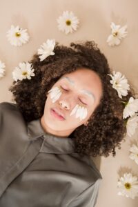 The Visual Fast: A Sensory Reset for Soul-Deep Rest | Calmly Rooted A person with curly hair lying down with eyes closed, surrounded by white daisy flowers, featuring flower petals placed delicately over their eyes.