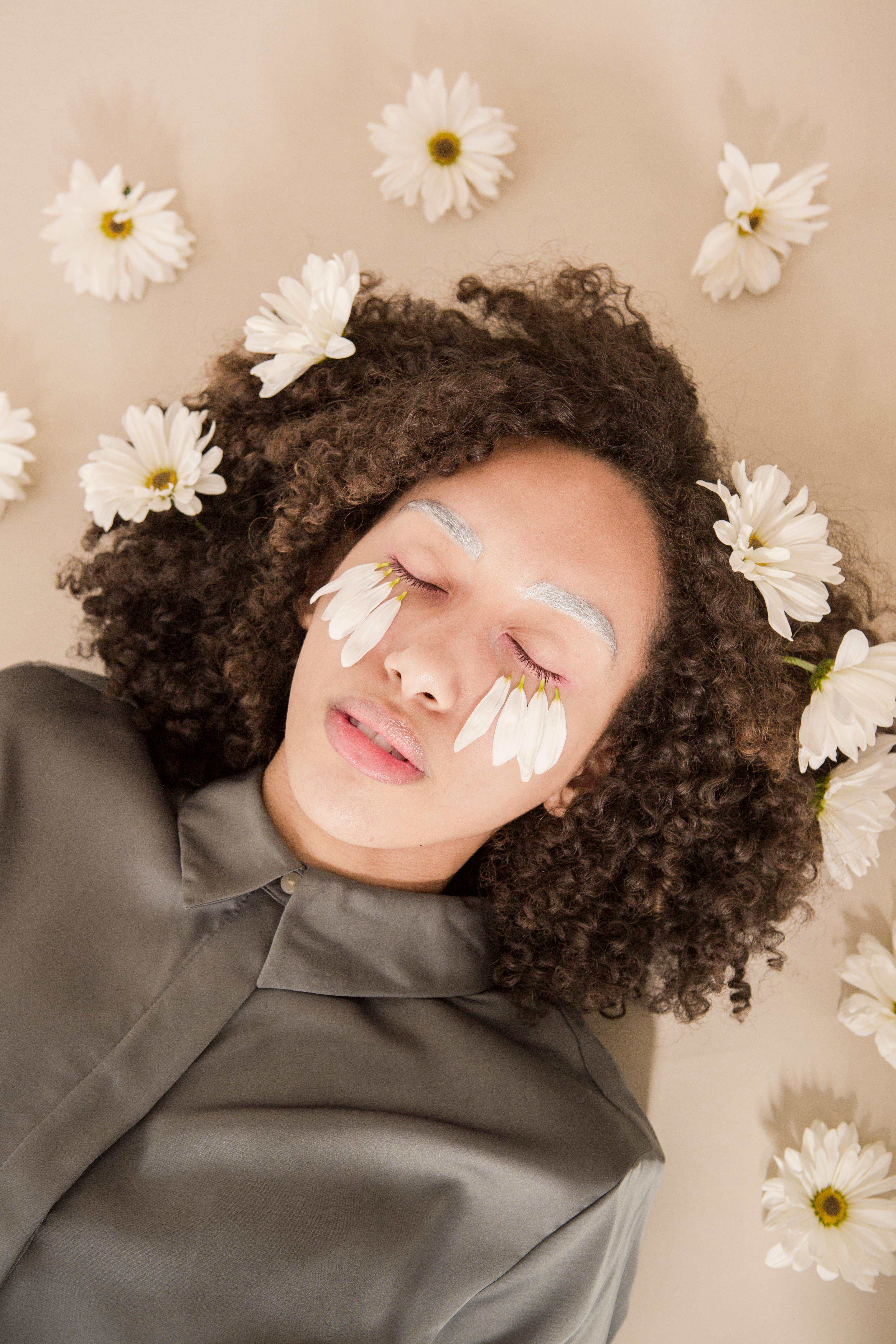 The Visual Fast: A Sensory Reset for Soul-Deep Rest | Calmly Rooted A person with curly hair lying down with eyes closed, surrounded by white daisy flowers, featuring flower petals placed delicately over their eyes.