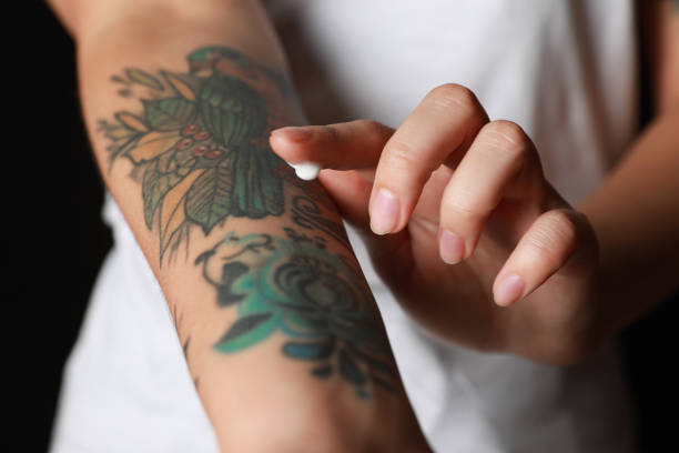 Woman applying cream on her arm with tattoos against black background, closeup | Calmly Rooted Woman applying cream on her arm with tattoos against black background, closeup