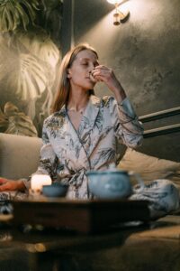 nightly-sleep-hygiene-recovery-ritual-calmly-rooted.jpg | Calmly Rooted A woman in a floral silk robe practicing a nightly tea ritual for sleep hygiene and recovery in a dimly lit, tranquil room with botanical wallpaper.