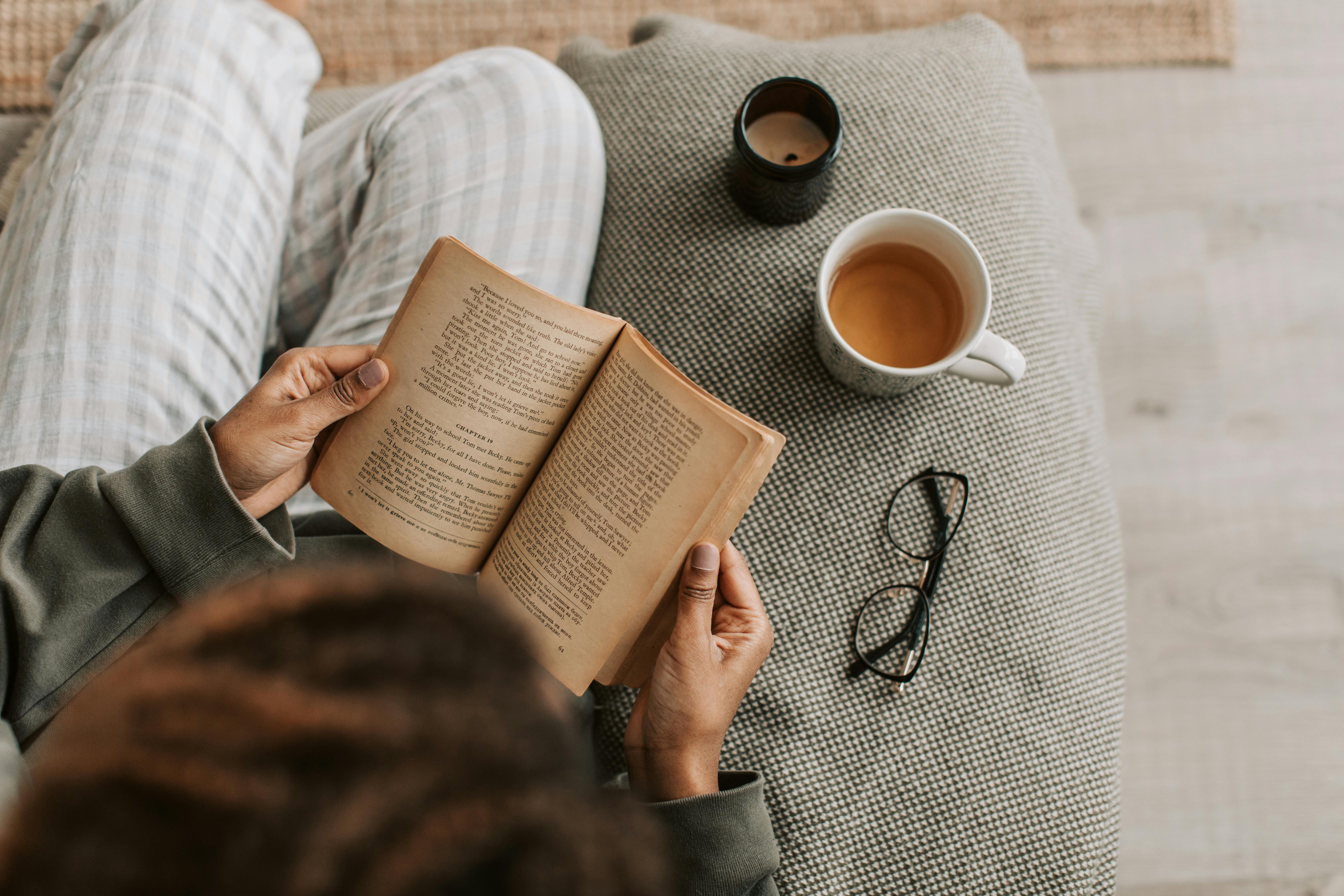 The Ultimate Evening Wind-Down for Better REM Sleep | Calmly Rooted A person relaxing on a textured sofa reading a book next to a warm cup of herbal tea and a candle, practicing a calming evening wind-down ritual.