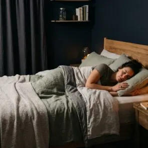 Bedroom Biohacking: Sensory Sleep Sanctuary | Calmly Rooted A woman sleeping peacefully in a dark, calming room optimized through bedroom biohacking, featuring warm low light on a clutter-free wooden nightstand, breathable sage green linen bedding, and an air purifier.