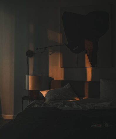Delayed Sleep Phase Syndrome Late Night | Calmly Rooted A dark, moody bedroom illuminated only by a dim, warm light casting shadows over an unmade bed, representing the late-night isolation of delayed sleep phase syndrome.