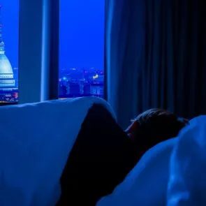 Functional Mushrooms for Sleep Calm Mind | Calmly Rooted A person lying in bed at night in a dark room overlooking a city, representing the struggle of a racing mind and the need for functional mushrooms for sleep.
