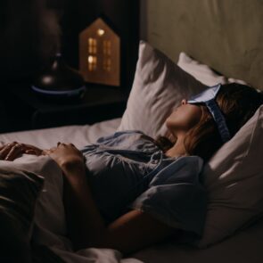 Restorative Bedtime Routine with Natural Sleep Aids and Diffuser | Calmly Rooted A woman sleeping peacefully wearing a light blue sleep mask, with a soothing essential oil diffuser and warm light on her bedside table, illustrating natural sleep aids for adults.