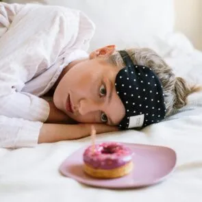 vanquish-blood-sugar-sleep-tips | Calmly Rooted A tired woman in bed with a sleep mask looking at a pink frosted donut, symbolizing how blood sugar and sugar cravings can disrupt sleep patterns.