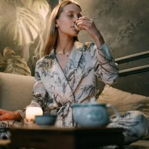 nightly-sleep-hygiene-recovery-ritual-calmly-rooted.jpg | Calmly Rooted A woman in a floral silk robe practicing a nightly tea ritual for sleep hygiene and recovery in a dimly lit, tranquil room with botanical wallpaper.