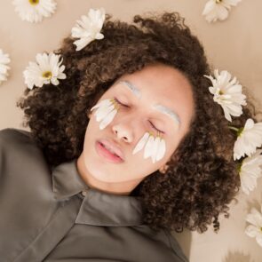 The Visual Fast: A Sensory Reset for Soul-Deep Rest | Calmly Rooted A person with curly hair lying down with eyes closed, surrounded by white daisy flowers, featuring flower petals placed delicately over their eyes.