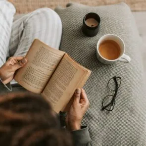 The Ultimate Evening Wind-Down for Better REM Sleep | Calmly Rooted A person relaxing on a textured sofa reading a book next to a warm cup of herbal tea and a candle, practicing a calming evening wind-down ritual.