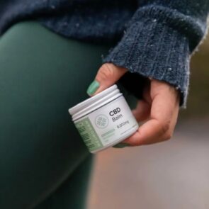 A close-up of a person wearing a cozy green sweater and green leggings holding a jar of extra strength 8000mg CBD relief balm against their leg.