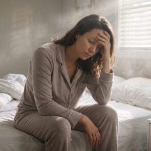 An exhausted woman sits on the edge of her unmade bed, head in hands, struggling to wake up. This image illustrates morning grogginess and the heavy feeling of sleep inertia as natural daylight filters through the room.