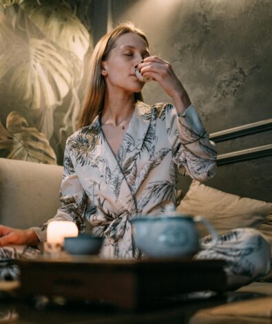 A woman in a floral silk robe practicing a nightly tea ritual for sleep hygiene and recovery in a dimly lit, tranquil room with botanical wallpaper.