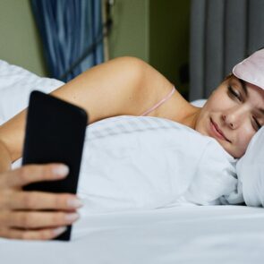 Woman in bed with a sleep mask checking her phone for deep sleep data and tracker results.