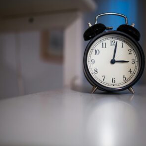 A black vintage-style twin-bell alarm clock sitting on a white surface in a dimly lit room, with the clock hands pointing to 3:00.