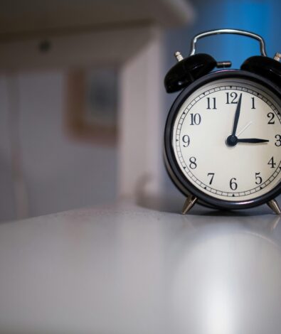 A black vintage-style twin-bell alarm clock sitting on a white surface in a dimly lit room, with the clock hands pointing to 3:00.