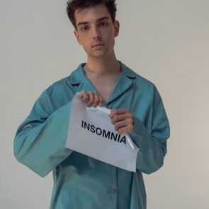 Tearing Down the Night: A Defiant Act to Banish Insomnia | Calmly Rooted A young person with dark curly hair and a neutral expression, wearing teal satin pajamas, looks directly at the camera while tearing a white piece of paper in half. The paper is printed with the word "INSOMNIA" in black capital letters.