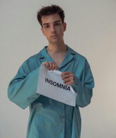 A young person with dark curly hair and a neutral expression, wearing teal satin pajamas, looks directly at the camera while tearing a white piece of paper in half. The paper is printed with the word "INSOMNIA" in black capital letters.