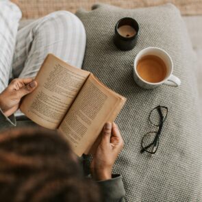A person relaxing on a textured sofa reading a book next to a warm cup of herbal tea and a candle, practicing a calming evening wind-down ritual.