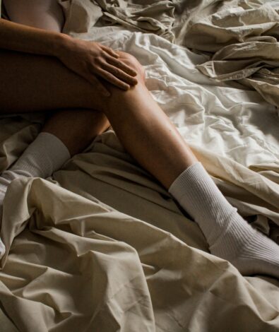 A person lying in bed with messy, tangled sheets in dim lighting, resting their hand on their knee in frustration due to restless legs at night.