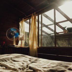A rumpled bed in a warm room with a desk fan running and bright sunlight streaming through the windows, representing the struggle of sleeping hot.