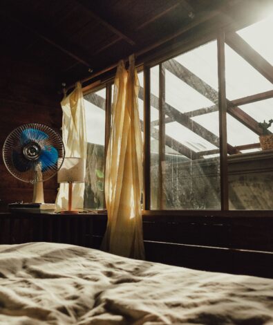 A rumpled bed in a warm room with a desk fan running and bright sunlight streaming through the windows, representing the struggle of sleeping hot.